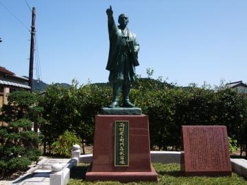 右手人差し指を上に指差している西村彦左衛門銅像の写真